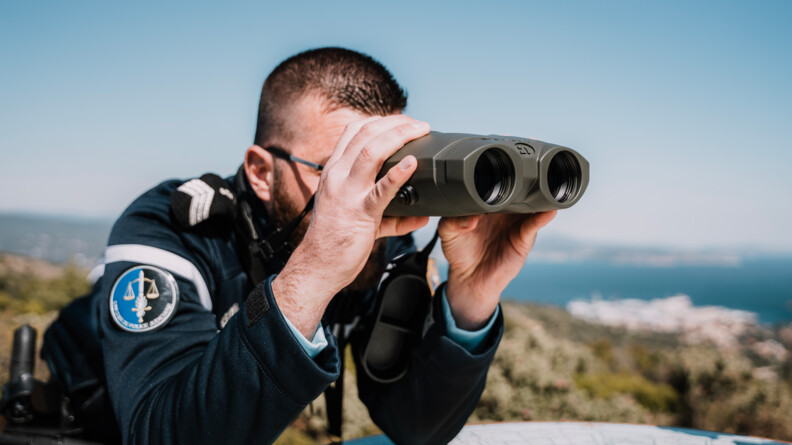 Un gendarme, accoudé à une table d'orientation, dans un site naturel verdoyant, en bordure de mer, examine un point au travers de jumelles télémétriques qu'il tient à deux mains, devant ses yeux.