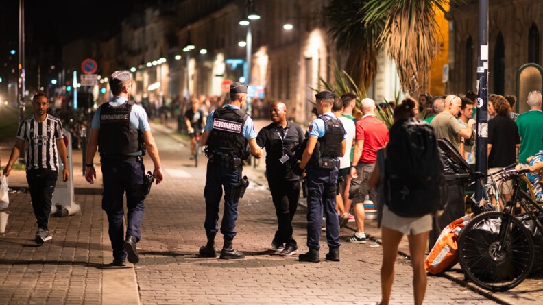 Trois gendarmes, vus de dos, en patrouille nocturne dans le centre-ville de Bordeaux où des gens se promènent. L'un d'eux échange avec un homme.