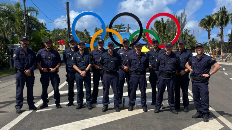 Une douzaine de militaires, vêtus du même uniforme bleu marine, se tiennent debout, côte à côte, devant les anneaux olympiques. Ils sont sur la route. le ciel est bleu, et l'on aperçoit des arbres, en particulier des palmiers, derrière eux.