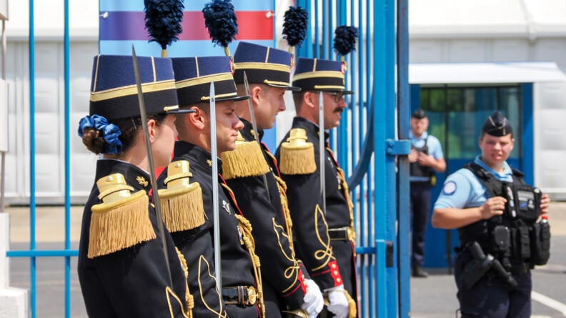 Quatre élèves-officiers de la gendarmerie en tenue de tradition, sabre à la main le long du corps, devant une grille bleue. A droite, flou, un gendarme en tenue d'intervention
