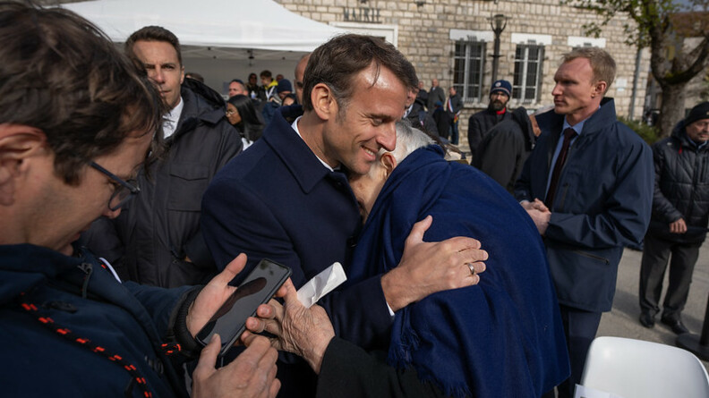 Agrandir l'image Le chef de l'Etat, Emmanuel Macron, tient dans ses bras, souriant, un vieil hmme recouvert d'une grande écharpe bleue, dissimulant son visage contre la poitrine du président.