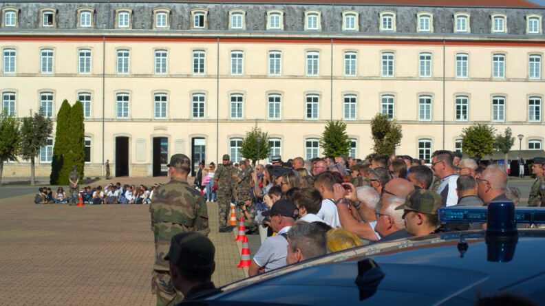 Devant un grand bâtiment de couleur clair se trouvent des jeunes assis par terre. A droite se trouve du public qui regarde dans la même direction. En tout premier plan se trouve le toit d'une voiture de gendarmerie avec un girophare étient. De dos se tient debout un militaire en tenue kakie