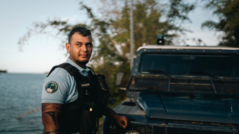 un gendarme adjoint volontaire de la gendarmerie &agrave; gauche de la photo, la main gauche pos&eacute; sur l'avant d'un v&eacute;hicule, regarde l&eacute;g&eacute;rement &agrave; gauche. Derri&egrave;re &agrave; gauche, un plan d'eau et &agrave; droite de la v&eacute;g&eacute;tation