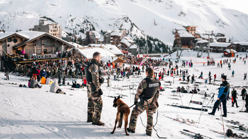 Agrandir l'image Deux gendarmes (un homme et une femme) du Groupe cynophile se tiennent debout, côte à côte, avec un malinois tenu en laisse. Les militaires se trouvent aux abord de la "Folie douce", établissement festif de la station d'Avoriaz, situé au bas des pistes enneigées, où sont regroupés de très nombreux touristes. venus faire la fête.