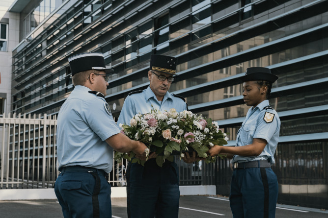 Trosi gendarmes, l'air grave, soutiennent une gerbe de fleurs, dans une cour.