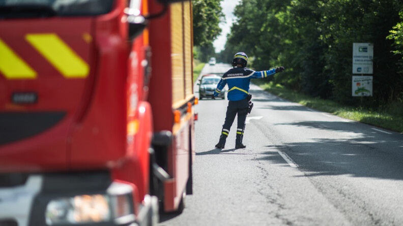 De dos sur la route un motocycliste de la gendarmerie est en train de faire la circulation. A gauche en flou un camion de pompier,