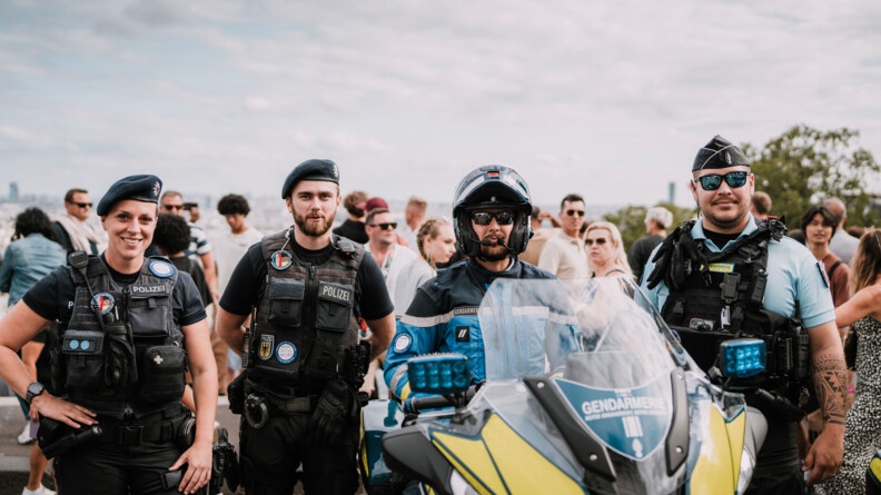 Deux policiers allemands, une femme, à gauche de l'image, et un homme, en tenue noire, sans manche, avec gilet pare-balles et béret sur la tête. La femme arbore un large sourire, une main sur la hanche, l'homme, est souriant, les mains derrière le dos. A leurs côtés, à droite, un motocycliste de la gendarmerie sur sa machine, casque sur la tête, visière relevée, lunettes de soleil sur le nez et micro devant la bouche. A côté de la moto, un second gendarme debout, de face, portant un calot, des lunettes de soleil, un gilet pare-balles par dessus un polo bleu clair. Derrière eux, une foule de passants anonymes.
