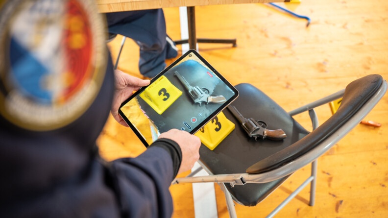 Agrandir l'image Vue sur les bras d'un homme portant une tablette qui photographie une revolver marqué d'un plot jaune portant le chiffre trois, posés sur une chaise