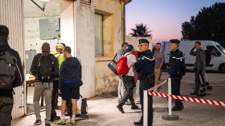 Devant un hangar, alors que le jour n'est pas totalement levé, des personnes font la queue pour faire vérifier leur badge auprès d'une militaire. Deux gendarmes de l'armement qui appuient leur collègue surveillent et échangent avec les habitués.