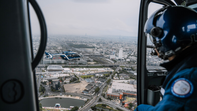 Vue prise du ciel, depuis l'habitacle d'un hélicoptère, d'un autre hélicoptère gendarmerie bleu en vol au-dessus d'une zone urbaine. Au premier plan, de profil, le mécanicien de bord, portant un casque, avec sur le bras la ronche JOP du ministère de l'Intérieur, est assis devant la porte latérale ouverte et observe l'autre appareil.