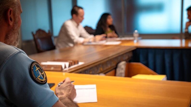 Agrandir l'image AU premier plan le bras d'un gendarme arborant une rondache présentant la balance de la Justice, derrière une table. Au fond flou, trois personnes autour d'une table