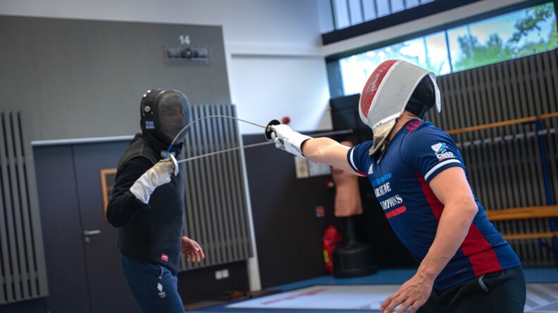 Un escrimeur vétu du survêtement de l'armée de champions touche son maître d'arme face à lui, vêtu de noir, qui ne pare pas