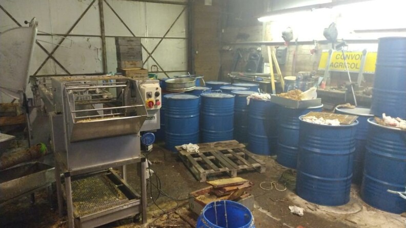 A droite une quinzaine de tonneaux m&eacute;talliques bleus et du mat&eacute;riel d'apiculture dans un hangar
