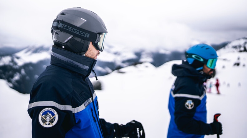 Agrandir l'image Deux gendarmes en ski se tiennent debout, de biais, en haut des pistes enneigées. Le ciel est gris et nuageux.