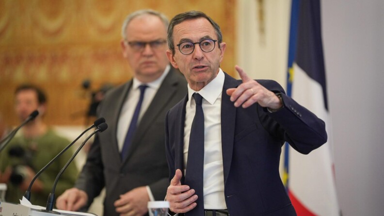Agrandir l'image Portrait en buste de Bruno Retailleau, en costume bleu marine, chemise blanche, cravate, pointant la main gauche devant lui. Au second plan, flou, François-Noël Buffet, ministre auprès du ministre d’État.