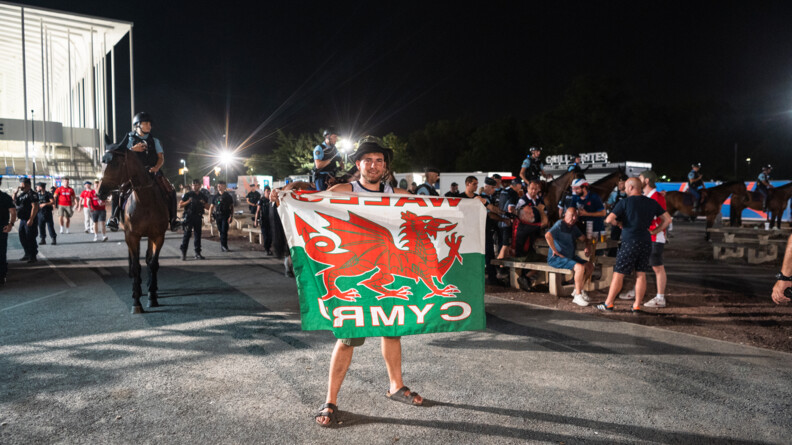 Au premier plan, un supporter gallois brandit un drapeau du Pays de Galles. Derri&egrave;re lui, d'autres supporters sont debout ou assis &agrave; des tables sur le parvis du stade, tandis que des cavaliers de la Garde, en ligne, suivis par des gendarmes mobiles &agrave; pied, avancent pour inciter les supporters &agrave; gagner les transports en commun ou le parking.