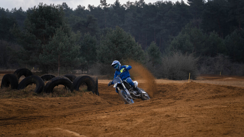 Un motard de la gendarmerie sur une moto cross bleue effectue un tournant dans un virage de sable sur un circuit d'entraînement; à gauche, à l'intérieur du virage, des pneux. EN arrière-plan, une forêt de résineux