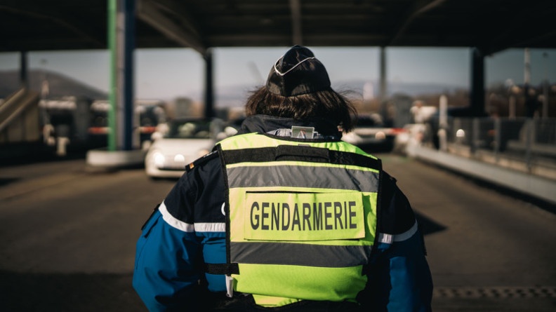 Agrandir l'image Image d'une femme gendarme, photographiée de dos, portant un gilet jaune portant l'inscription "Gendarmerie", tournée vers la gare de péage de l'autoroute. Une voiture blanche avance dans sa direction.