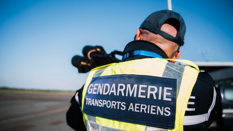 Agrandir l'image Sur le tarmac d'un aéroport, un gendarme avec un gilet jaune sur lequel est indiqué "Gendarmerie Transports aériens" pointe un fusil anti-drone vers le ciel.
