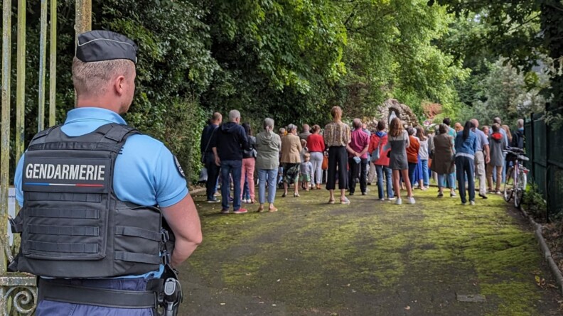 Agrandir l'image En extérieur, un gendarme en premier plan se tient debout et surveillent une cérémonie religieuse qui se déroule en second plan.