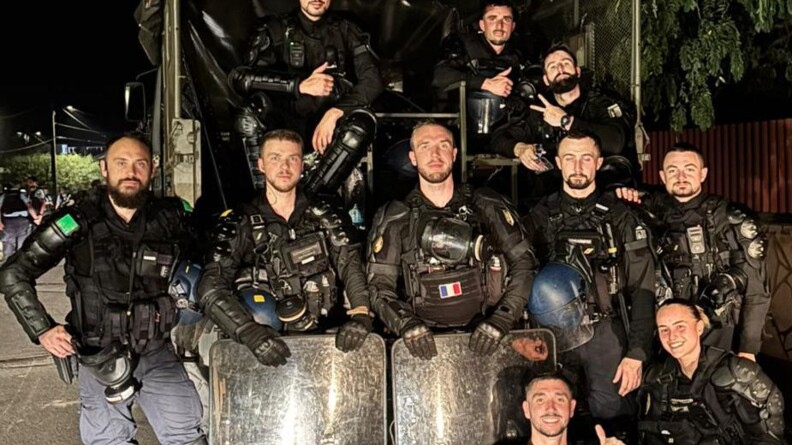 Dix gendarmes mobiles en tenue de maintien de l'ordre, sans casque, à l'arrière d'un véhicule TRM. Trois sont assis à l'arrière du véhicule, cinq sont debout devant eux, dont deux derrière leur bouclier, et deux autres sont accroupis au premier plan.