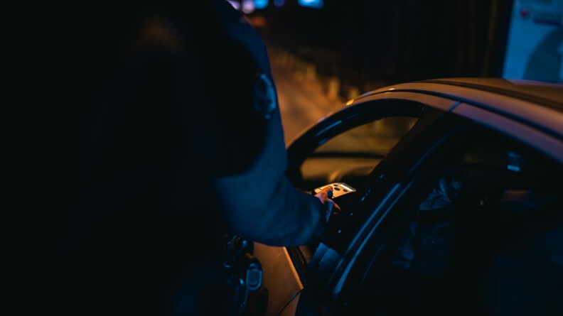 Un gendarme de dos. Il tient dans sa main un éthylotest qu'il est en train de tendre à un conducteur. Il fait nuit et sombre.