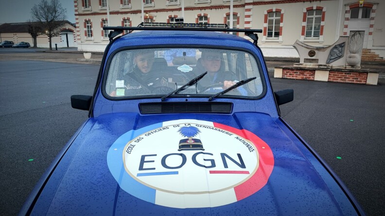 Deux hommes roulent &agrave; bord d'une Renaud 4L bleu marine, dont le capot est couvert d'un grand &eacute;cusson comportant la mention "Ecole des officiers de la gendarmerie nationale - EOGN". Ils circulent dans l'enceinte d'un b&acirc;timent, que l'on aper&ccedil;oit en arri&egrave;re plan. Des gouttes de pluie sont visibles sur le capot de la voiture.