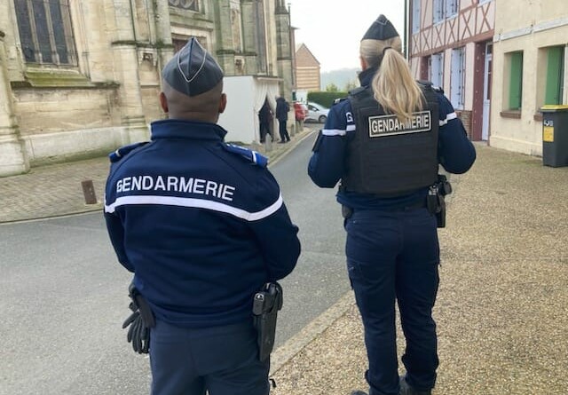 Deux gendarmes de dos au premier plan, à gauche un homme et à droite une femme, dans une rue. A gauche de la rue le mur d'un église, à droite la façade d'un bâtiment normand