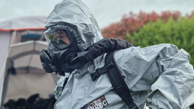 Un homme en combinaison grise, portant sur la poitrine le signe F2NRBC, ainsi qu'un masque sur son visage, avance dans un espace extérieur