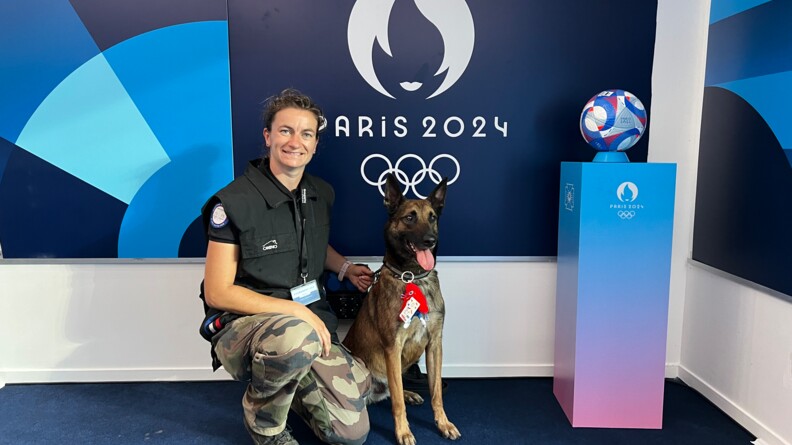 Photo d'une femme gendarme, accroupie, souriante, posant aux c&ocirc;t&eacute;s de son malinois assis &agrave; sa gauche. Derri&egrave;re eux, une affiche sur fond bleu marine porte l'inscription"Paris 2024"