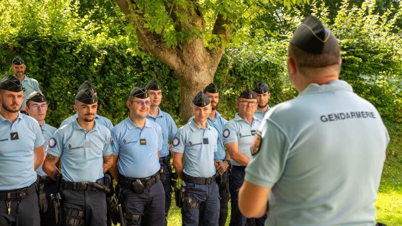 Plusieurs gendarmes en uniforme se tiennent debout, côte à côte, dans un espace extérieur verdoyant, face à leur supérieur, que l'on aperçoit de dos