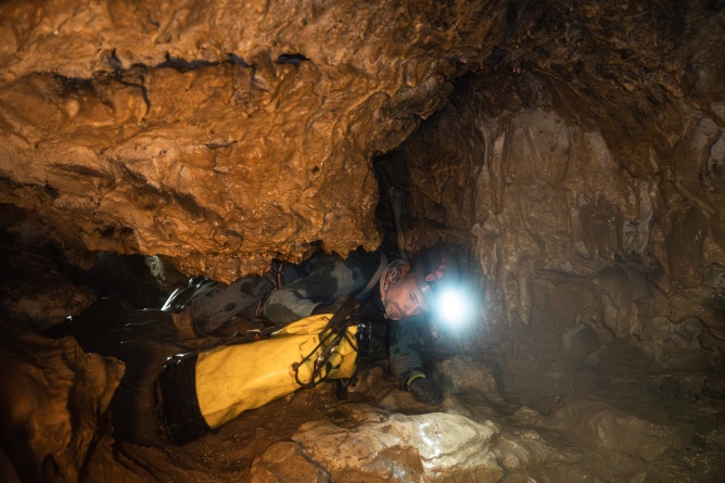 Agrandir l'image Photo montrant un gendarme spéléologue en combinaison et casque équipé d’une lampe frontale, en train de sortir d'un boyau étroit à plat ventre, poussant un sac jaune devant lui, à l’intérieur d’une grotte.