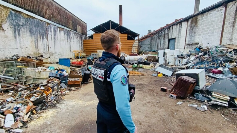 Un gendarme se tient debout, de dos, dans une cour, dont le sol est jonché de déchets.