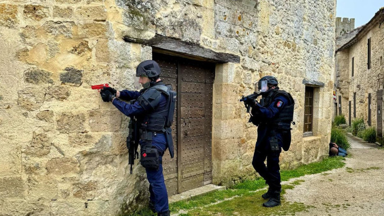 Deux gendarmes équipés en gilet lourd et avec des armes fictives rouge arrivent à un coin de mur