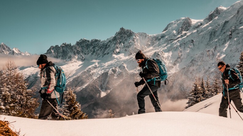 Trois randonneurs en raquettes sur la neige, vus de profil, avec la forêt et la montagne enneigées en fond.
