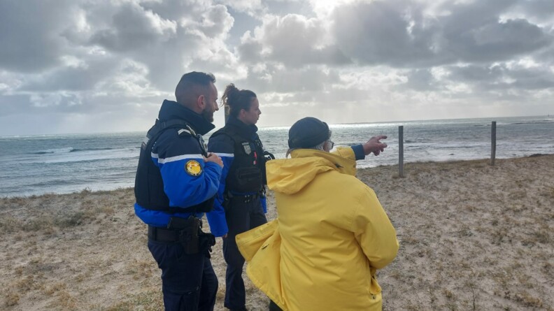 Deux gendarmes, un homme et une femme, se tiennent debout, de biais, en bordure de plage. Ils échangent avec une personne vêtue d'un ciret jaune qui pointe l'horizon du doigt. Tous trois se tiennent sur un sol sablonneux. Derrière eux, on aperçoit la mer. Le ciel est gris et nuageux.