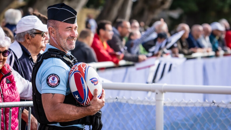 Devant les barrières sécuirsant un public, de profil droit et souriant, tenant dans son bras droit un ballon de la coupe du monde siglé CHili