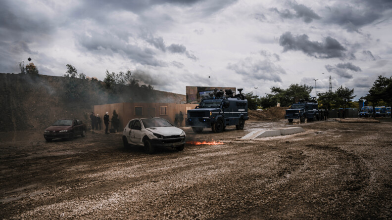 Point de vue global de la démonstration: à gauche deux véhicules épaves et quatre manifestants se dissimulant derrière un bâtiement. Deux centaure arrivent vers les véhicules, suivis par un peloton de gendarmes mobiles à pied. Des véhicules de maintien de l'ordre arrivent sur la droite