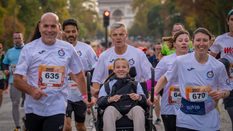 Des coureurs, vêtus d'un T-shirt blanc avec un numéro sur le ventre, courent sur une avenue pavée, en poussant le fauteuil roulant d'une jeune femme blonde, souriante, portant des tresses. On aperçoit l'arc de triomphe en arrière plan.