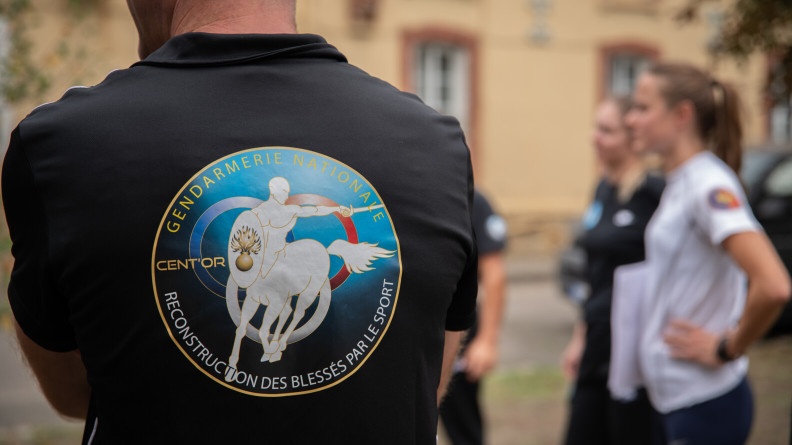 Agrandir l'image Un homme de dos, en t-shirt noir, avec un logo sur la largeur du dos représentant un centaure blanc, avec une épée et un bouclier portant une flamme. Sont inscrites les mentions : GENDARMERIE NATIONALE - CENT'OR et RECONSTRUCTION DES BLESSES PAR LE SPORT. A droite, au second plan, deux personnes de profil.