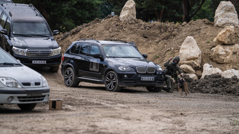 Devant deux 4x4 noirs, un ma&icirc;tre de chien du GIGN envoie son animal qui court enlui indiquant un point au laser