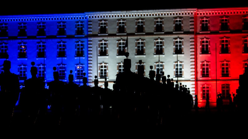 Agrandir l'image Vue en contre-plongée des élèves de l'EOGN, en silhouettes noires, devant l'état-major de l'Ecole illuminé de bleu blanc et rouge