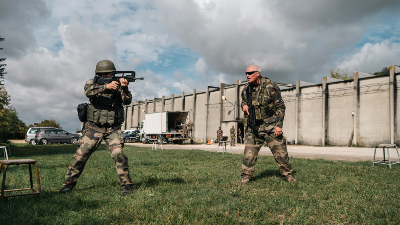 Au premier plan &agrave; gauche, un gendarme en tenue de combat pointant son famas en direction de l'avant-droite. A trois m&egrave;tres de lui &agrave; sa gauche, un instructeur en treillis, tenant un FAMAS dans la maint droite le long de son corps, l'invective. En arri&egrave;re-plan, de droite &agrave; gauche, les bat&icirc;ments en b&eacute;ton de Beynes, un camion banc en cours de d&eacute;chargement, d'autres militaires et quelques vehicules stationn&eacute;s