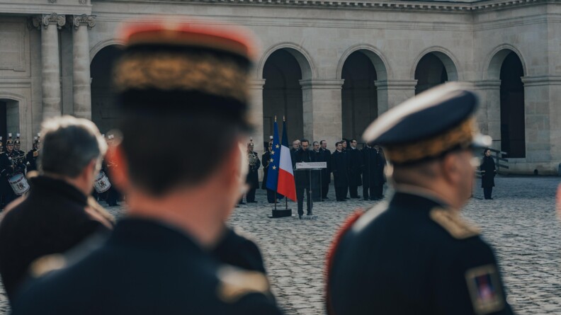 En flou au premier plan, un général de gendarmerie et le préfet de police, au fond le ministre de l'Interieur derrière un pupitre et à sa droite les drapeaux français et européens, dans la cour des invalides