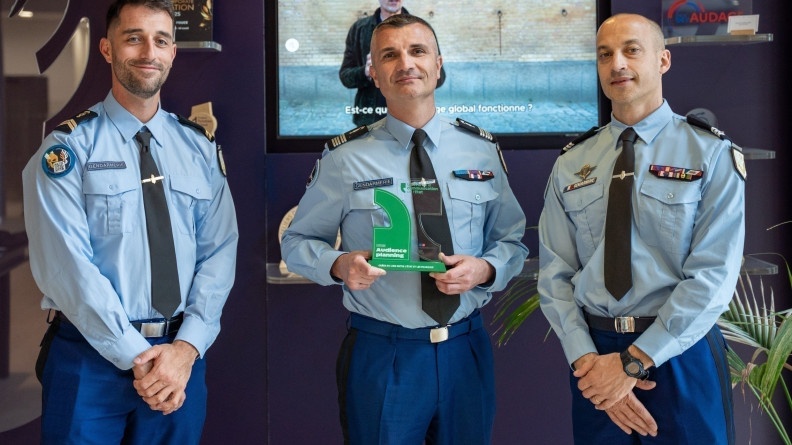 Trois gendarmes, debout, côte à  côte, posent souriants face à l'objectif. L'un d'eux (celui du milieu) tient dans ses mains un trophée de couleur verte