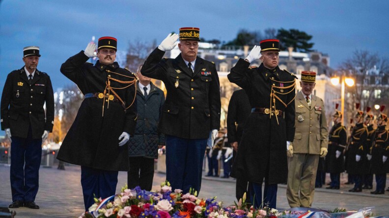 Au premier plan, un g&eacute;n&eacute;ral de gendarmerie, encadr&eacute; par deux gendarmes, tous trois en grande tenue et au garde-&agrave;-vous devant une gerbe de fleurs.