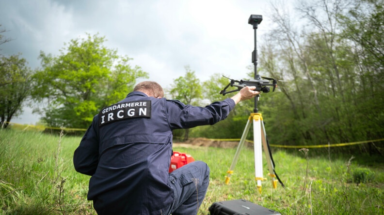 Un gendarme est accroupi dans une clairière. Il porte un drone avec sa main droite. Avec celui-ci, il va faire une reconnaissance de terrain.