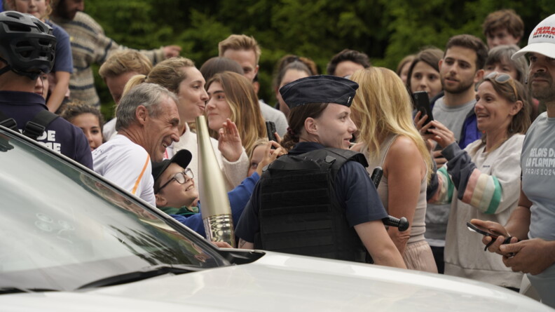 Derrière un véhicule, une gendarme mobile de profil, à sa gauche une homme portant la flamme olympique, le tout tèrs proche de la foule, tous prenant des photos et souriant