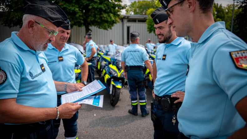 Qautre gendarmes se tenant debout dans une cour sont réunis. L''un d'eux parle, tout en regardant les papiers qu'il tient des papiers dans ses mains. En arrière plan, on paerçoit des motocyclettes de la gendarmerie, ainsi que des gendarmes se tenant à côté.