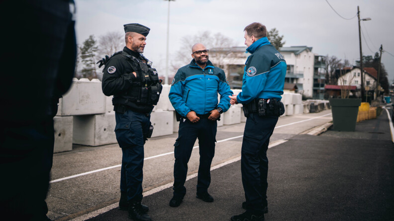 Agrandir l'image Un gendarme français discute avec deux policiers suisses. Les trois hommes se tiennent debout sur un trottoir. Deux sont de biais par rapport à l'objectif, tandis que le troisième homme est de face. Le temps est gris.
